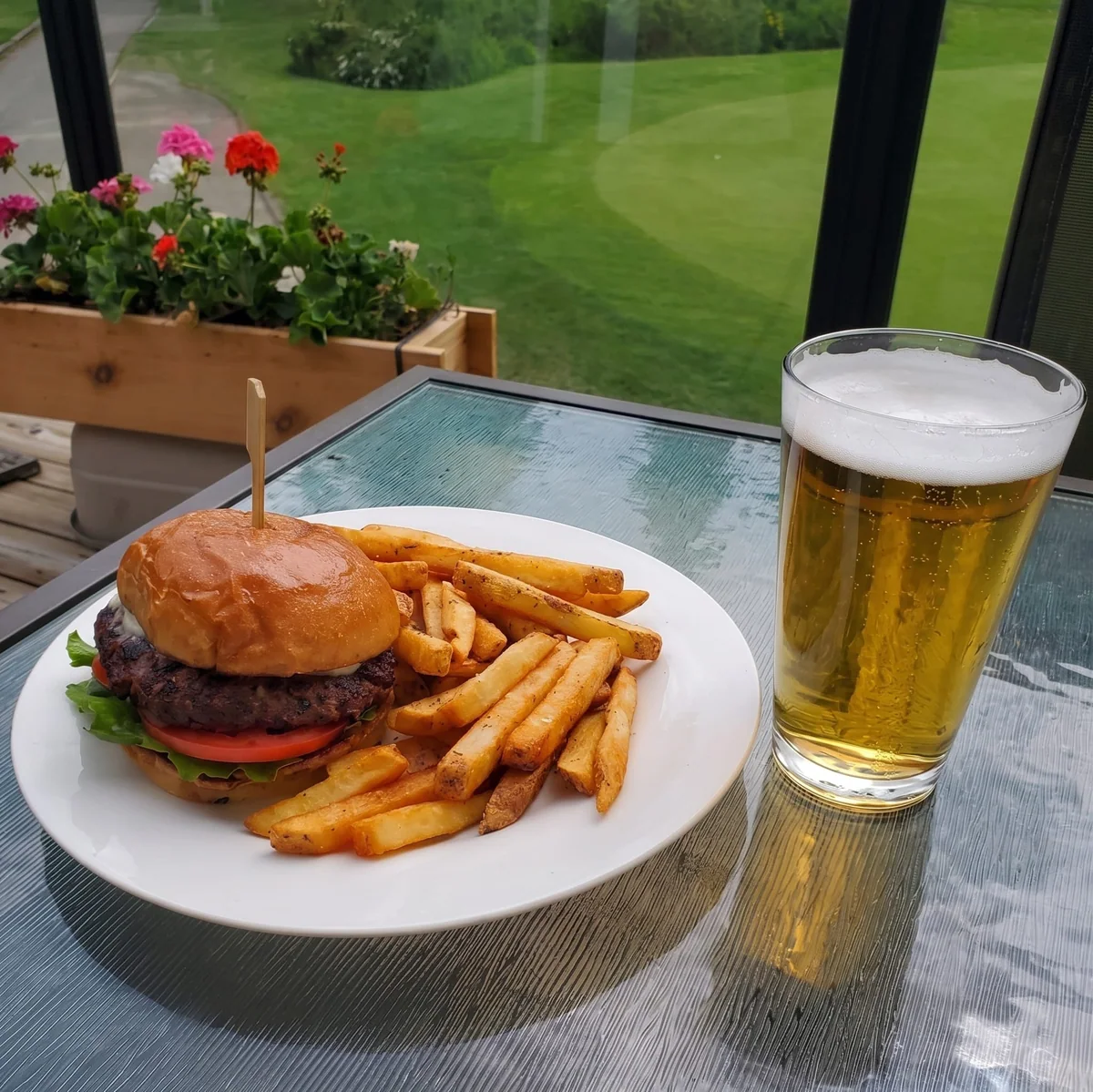 A burger and fries on a white plate beside a tall pint of beer, the course visible through the patio window