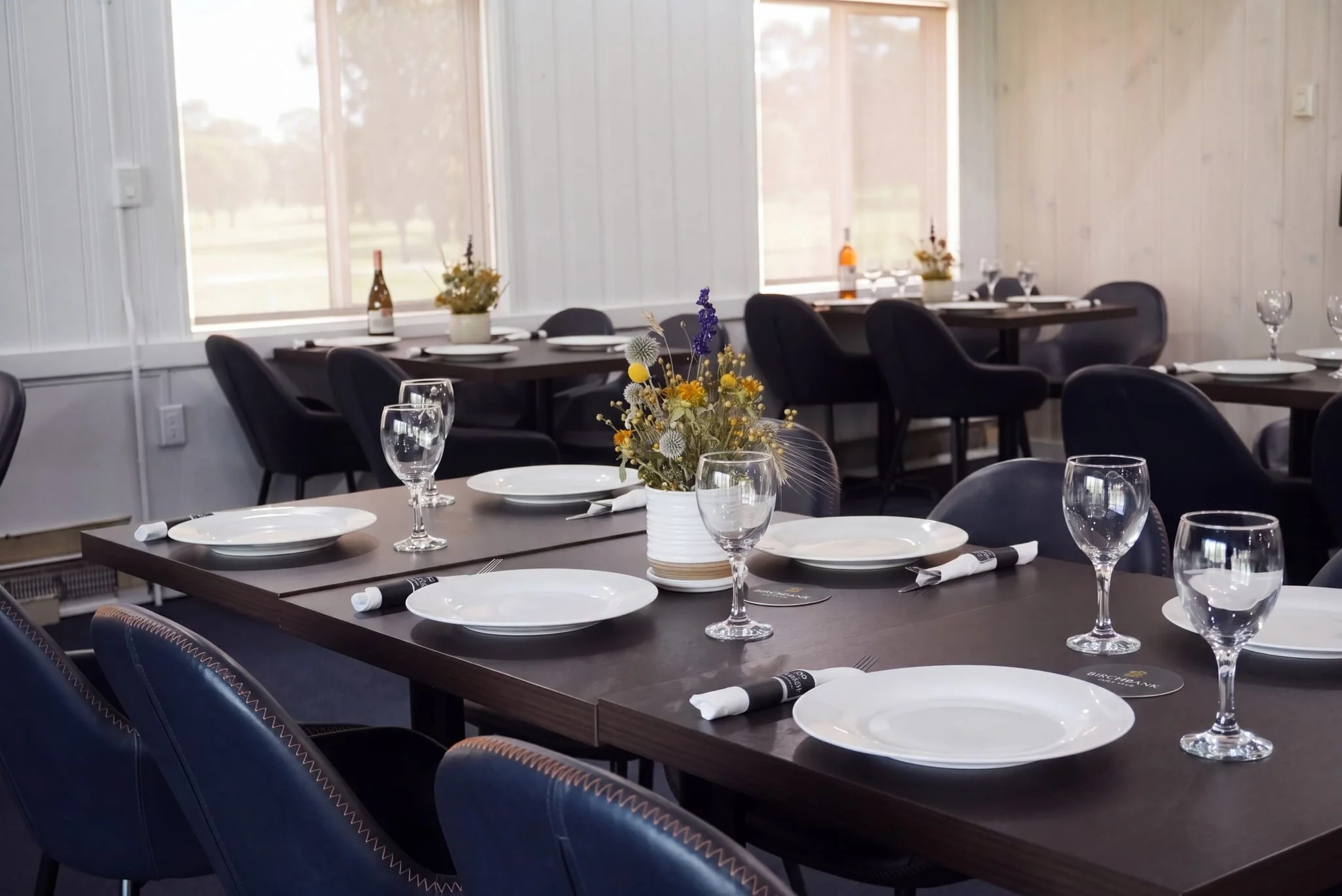 The Bistro dining room at Birchbank set with white linen, wine glasses, and dried-flower centrepieces, the course visible through the windows