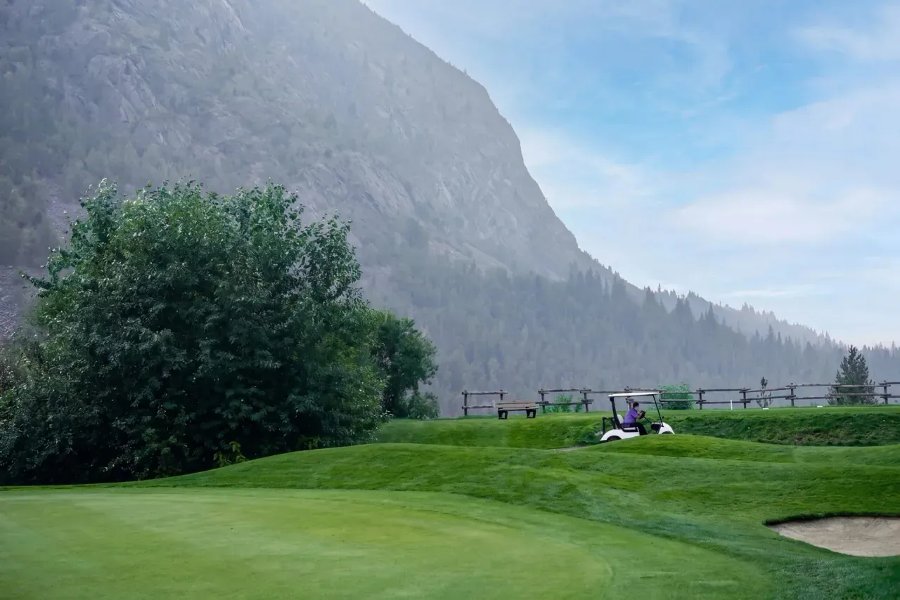 A single golf cart parked along the fence line at Birchbank with the Selkirk mountains rising behind