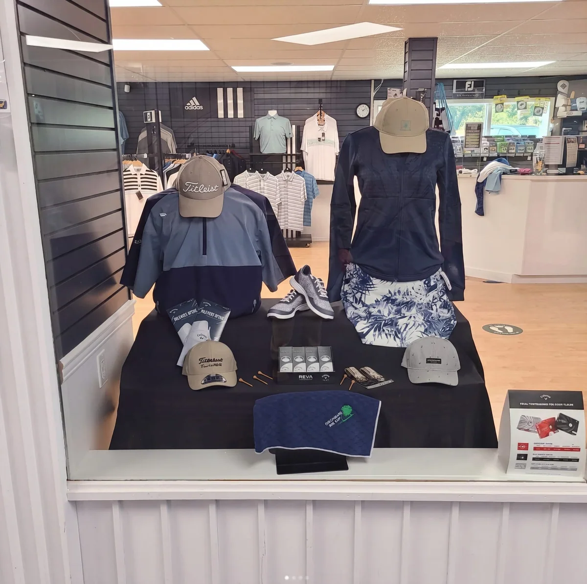 A staged Pro Shop apparel display: navy and grey Titleist hats, an adidas branded backdrop, FootJoy shoes on the front edge of the display, and Birchbank-branded golf accessories